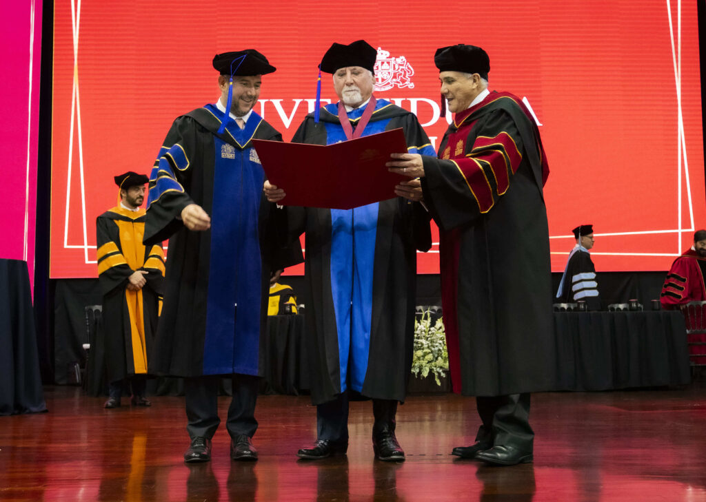 Autoridades de la Universidad San Pablo de Guatemala entregan el título Honoris Causa a Steve Moore durante la ceremonia de graduación 2026, sobre el escenario con togas académicas azules y negras