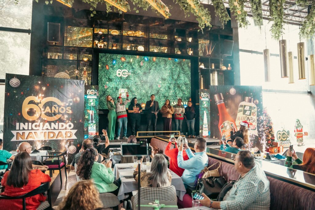 Evento 'B&B 60 años Iniciando Navidad' con invitados en el escenario y asistentes celebrando la temporada navideña